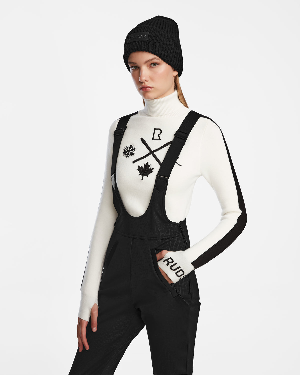 A woman wears the Rudsak OKSANA - 8124527 Monogram Print Black beanie, a white turtleneck with black graphics, and fitted flared ski pants, standing against a plain white background with a serious expression.