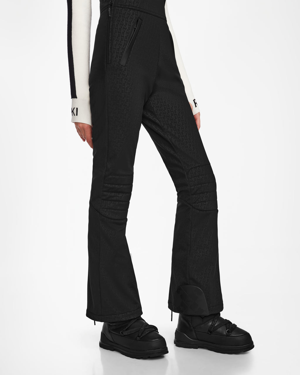A person wearing Rudsak OKSANA - 8124527 Monogram Print Black fitted flared women’s ski pants, black snow boots, and a white long-sleeve top with black striped cuffs featuring an “F,” standing against a white background.
