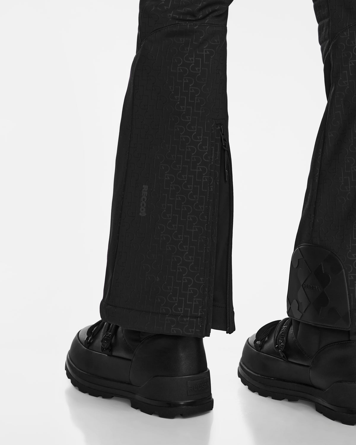 Close-up of a person wearing Rudsak OKSANA - 8124527 Monogram Print Black fitted flared ski pants with a partially open side zipper and black sneakers, standing on a white background.