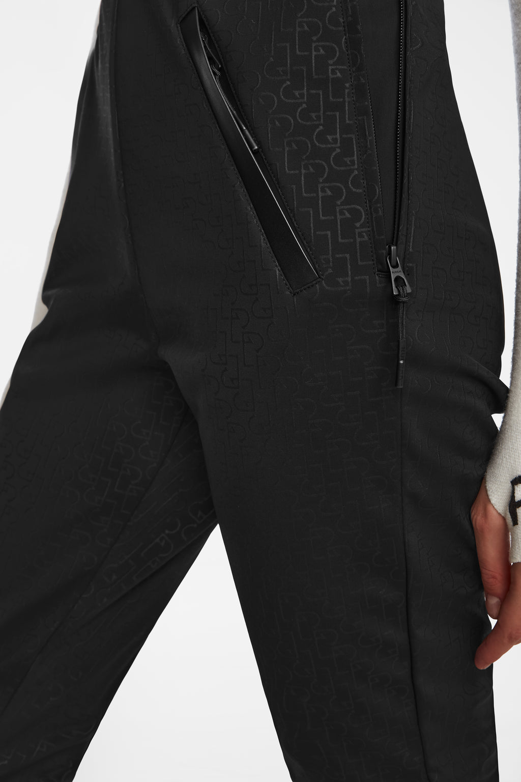 Close-up of a person wearing Rudsak OKSANA - 8124527 Monogram Print Black fitted flared ski pants with subtle embossed patterns and zippered pockets. Only the lower torso and hand are visible; paired with a white top with black sleeve text.