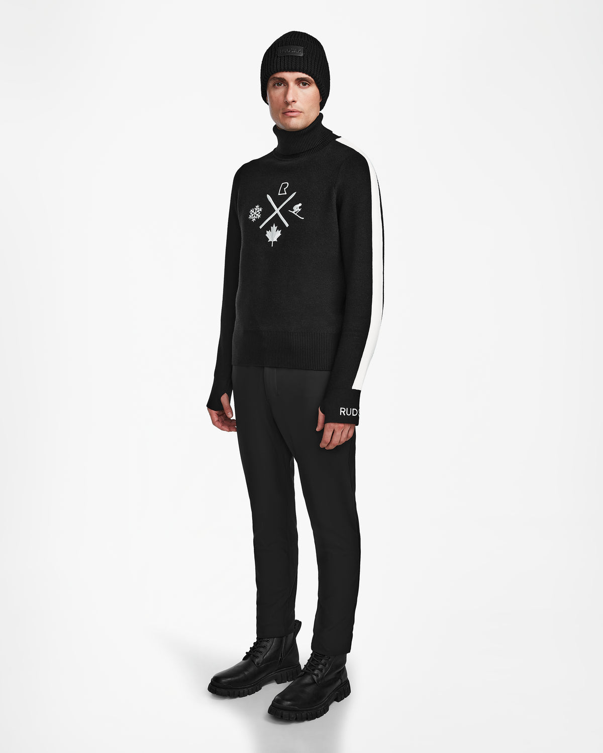 A person stands against a white background wearing Rudsaks OLIVIER - 6524545 Black mens ski turtleneck with white symbols—styled with a black beanie, black pants with a white side stripe, and black boots for an effortlessly cool winter look.
