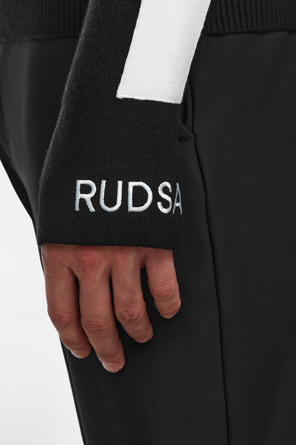 A close-up of a hand against black pants shows the sleeve of the OLIVIER - 6524545 Black après-ski sweater by Rudsak, with RUDSAK embroidered in white near the wrist—a stylish winter wardrobe essential.