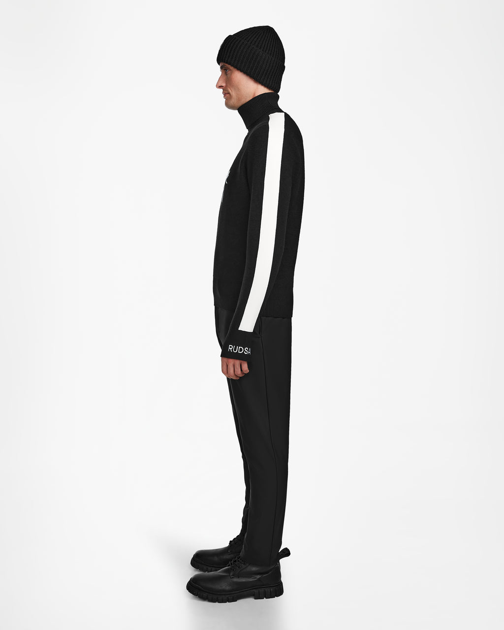 A person stands in profile wearing OLIVIER - 6524545 Black by Rudsak—a black beanie, black jacket with a white sleeve stripe, black pants, and boots—creating an effortlessly cool winter look against a plain white backdrop.