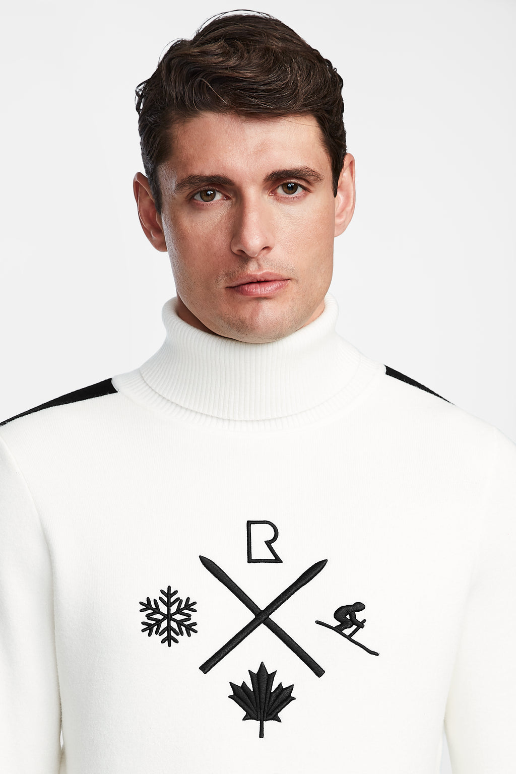 A man with short brown hair wears the Rudsak OLIVIER - 6524545 Optic White men’s ski turtleneck, featuring black icons—snowflake, crossed skis, maple leaf, skier, and R—ideal for winter wardrobes or après-ski events.