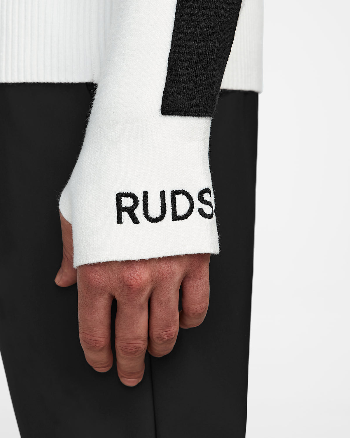 A close-up of a person wearing the Rudsak OLIVIER - 6524545 Optic White mens ski turtleneck sweater with a black stripe and embroidered logo near the cuff—paired with black pants, it’s a winter essential.