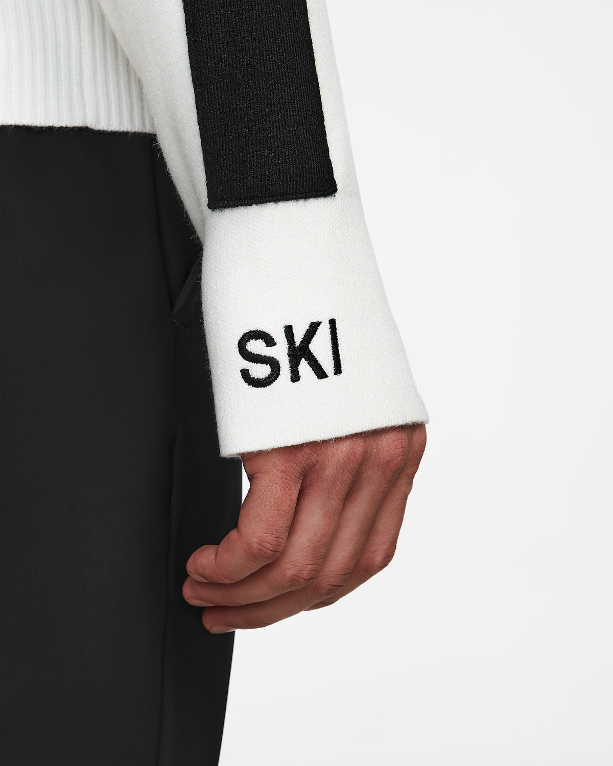 A close-up highlights a hand resting by the side, featuring the Rudsak OLIVIER - 6524545 Optic White sweater with SKI on the sleeve, paired with black pants—an effortlessly chic après-ski ensemble.