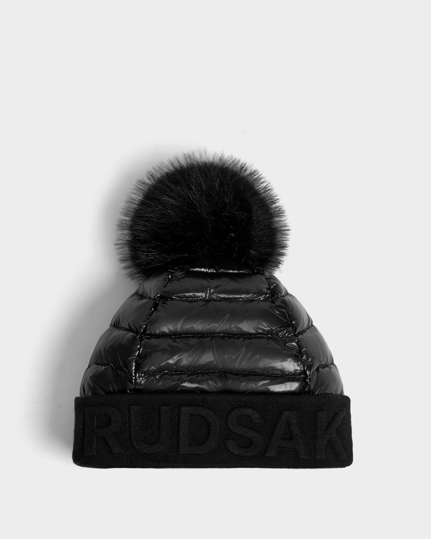 The OLYA - 8422504 BLACK by Rudsak is a black puffer-style women’s winter hat with a large fur pompom and wide cuff featuring bold RUDSAK embroidery, shown against a plain light gray background.