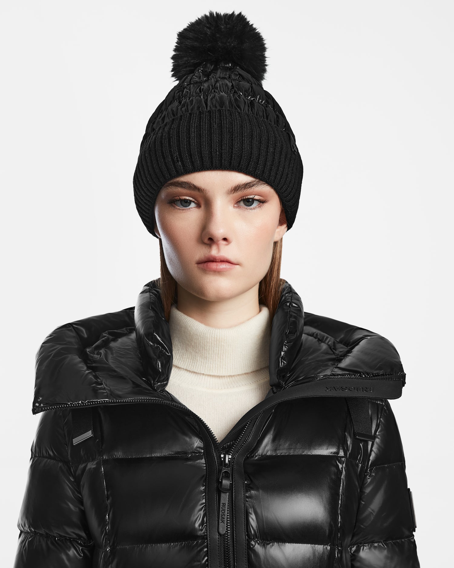 A woman wears the Rudsak OLYA - 8422504 BLACK winter hat with a fur pompom, a cream turtleneck, and a shiny black puffer jacket, looking directly at the camera against a white background.