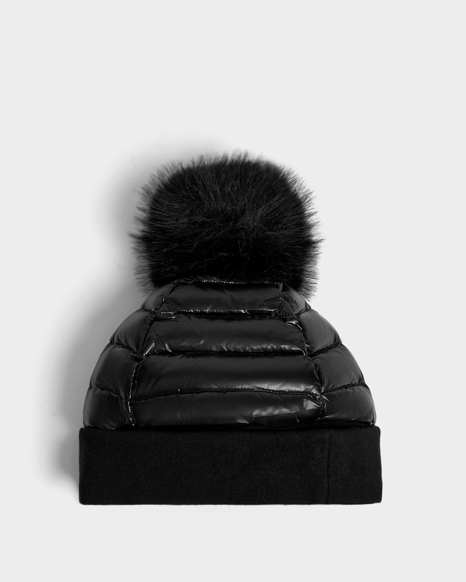 The Rudsak OLYA - 8422504 BLACK is a shiny black puffer beanie hat featuring a wide cuff and a large, fluffy fur pompom, shown against a light gray background.