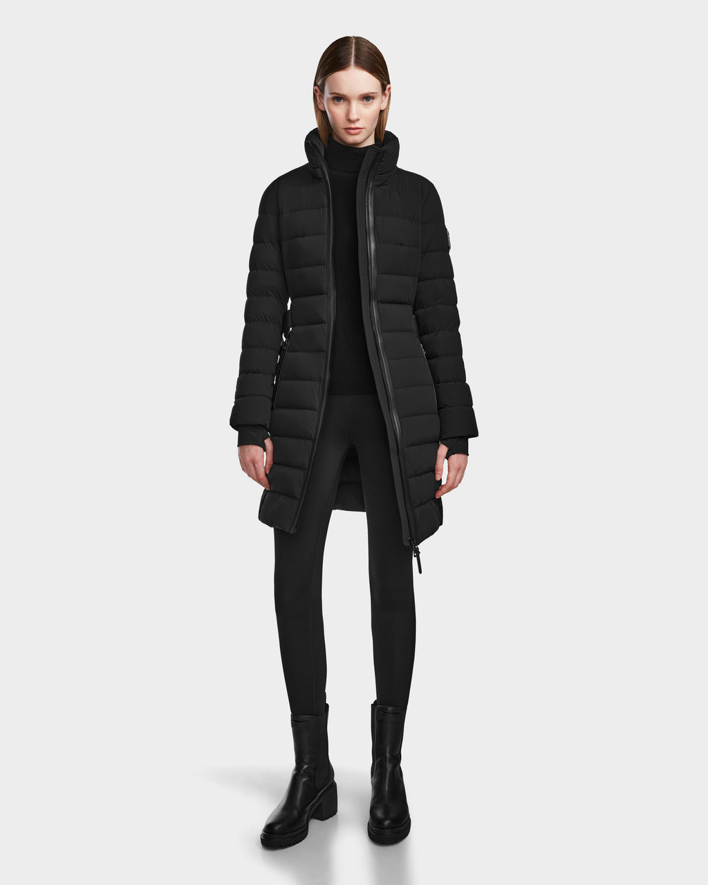 A woman faces forward against a white background, wearing the Rudsak PELIN M - 8124814 Black womens down puffer coat with a black turtleneck, leggings, and boots.