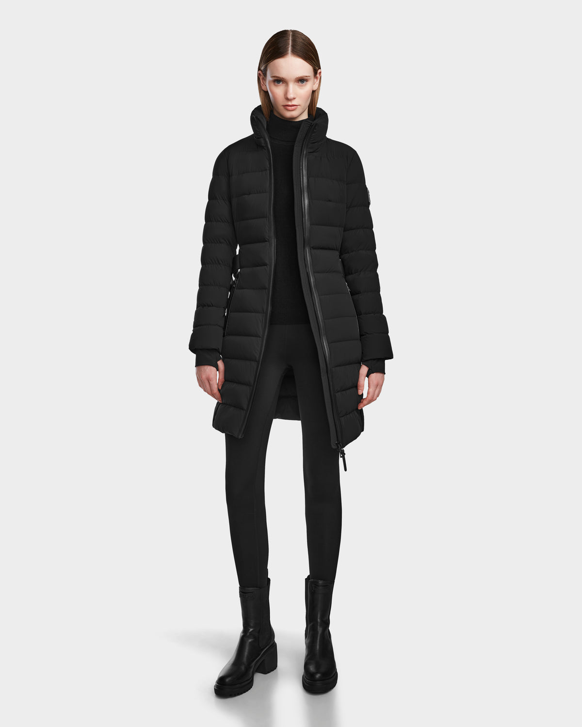 A woman faces forward against a white background, wearing the Rudsak PELIN M - 8124814 Black womens down puffer coat with a black turtleneck, leggings, and boots.