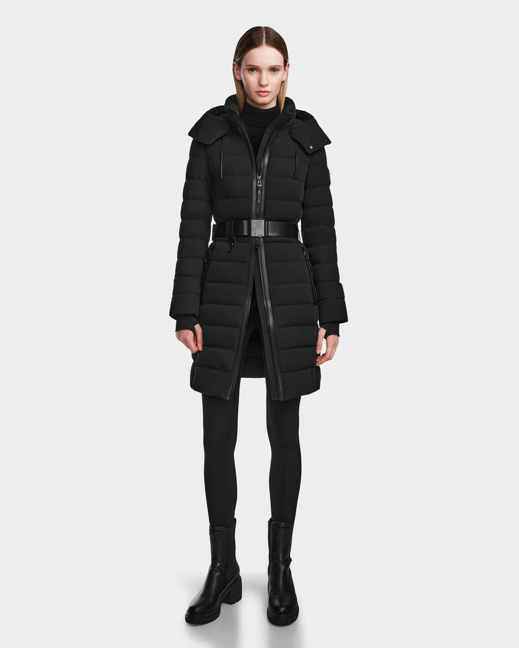 A woman faces forward wearing the Rudsak PELIN M - 8124814 Black belted knee-length down puffer with a hood, styled with black leggings and ankle boots, set against a plain white background.