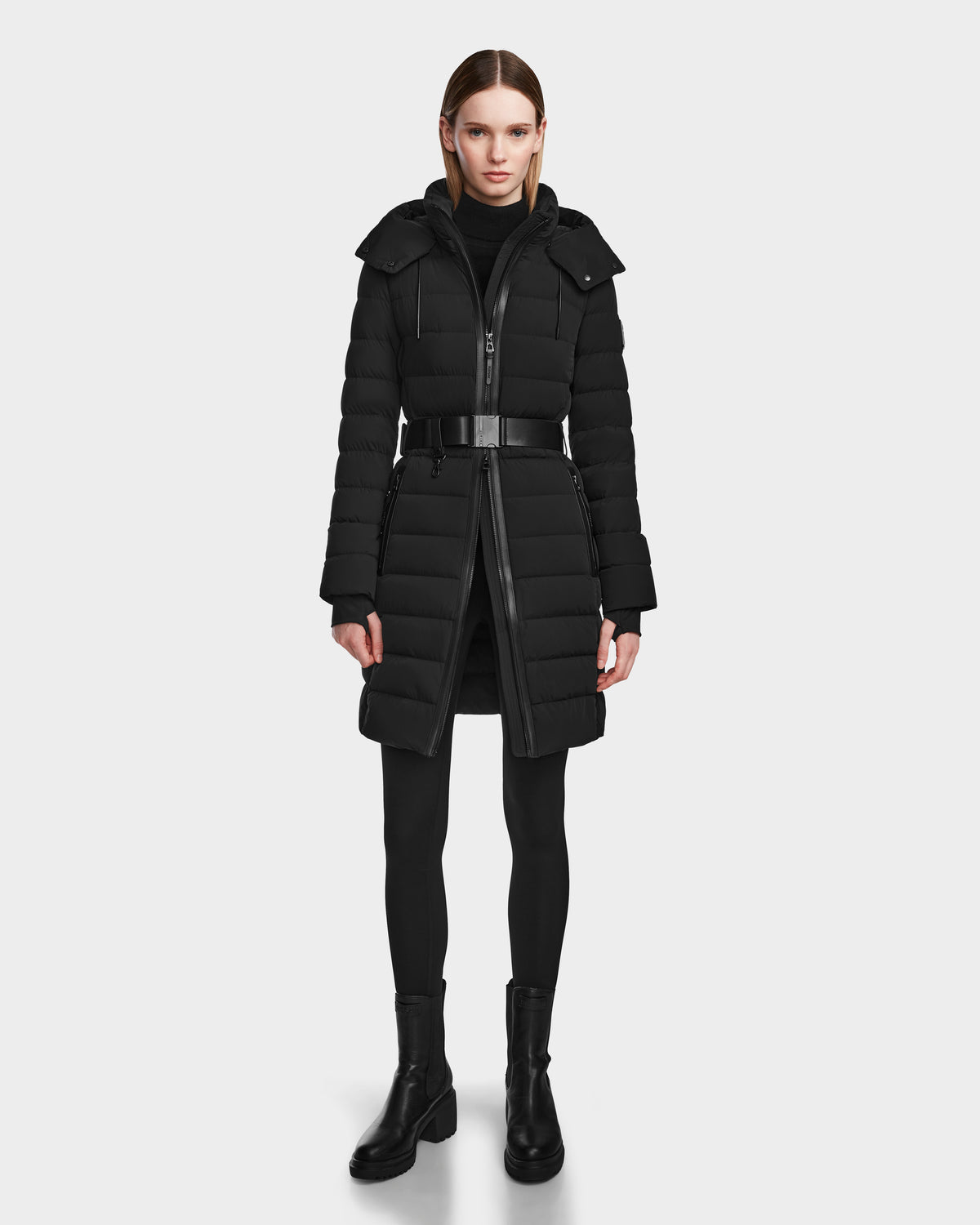A woman faces forward wearing the Rudsak PELIN M - 8124814 Black belted knee-length down puffer with a hood, styled with black leggings and ankle boots, set against a plain white background.