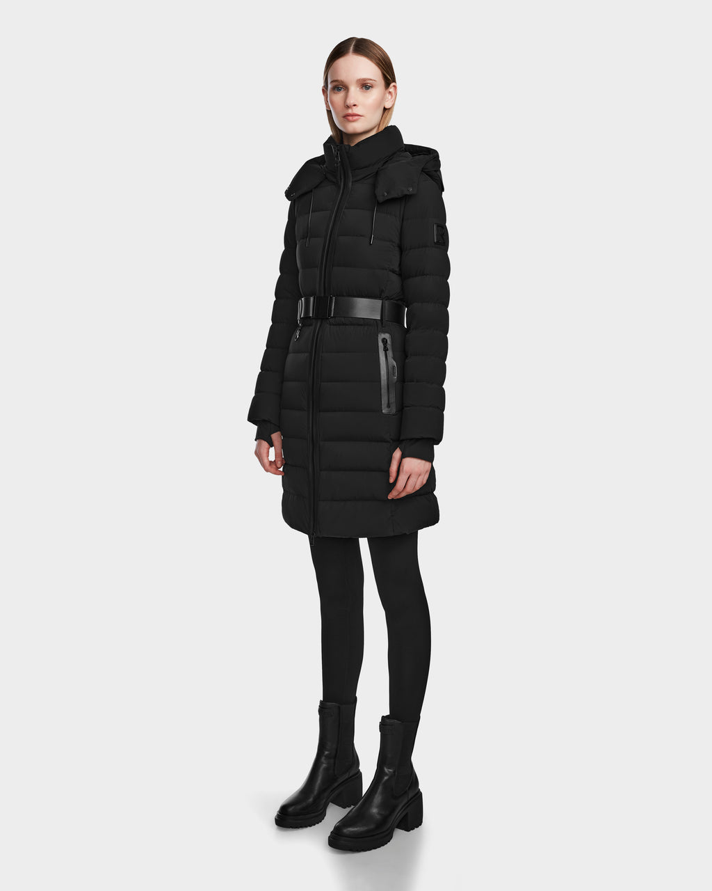 A woman wears the Rudsak PELIN M - 8124814 Black down puffer coat with a hood, paired with black leggings and ankle boots, standing against a plain white background.