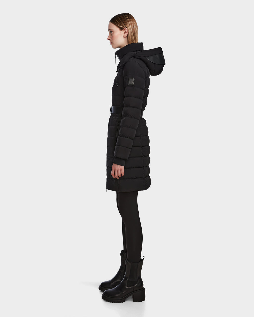A woman stands in profile wearing the Rudsak PELIN M - 8124814 Black down puffer coat with a belt and hood, paired with black leggings and ankle boots against a plain white background.