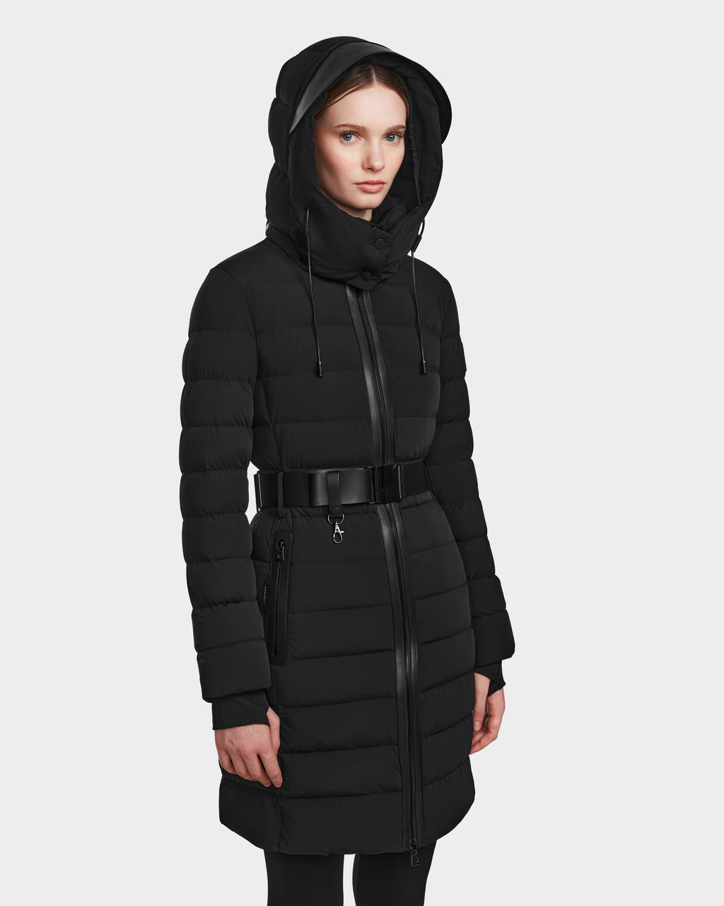 A woman models the Rudsak PELIN M - 8124814 Black, a hooded, belted, quilted jacket with a zippered front—ideal for effortless winter style—posed against a plain light background with her hands relaxed at her sides.