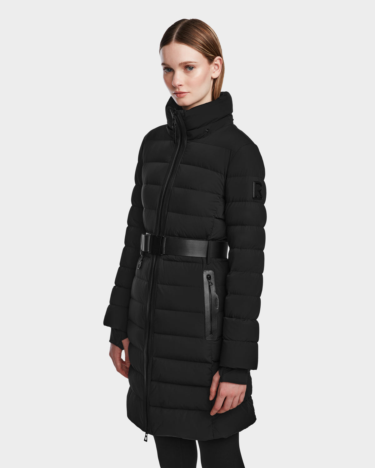 A woman wears the Rudsak PELIN M - 8124814 Black long down puffer coat with a belted waist, high collar, quilted design, and zippered pockets, standing against a plain white background.