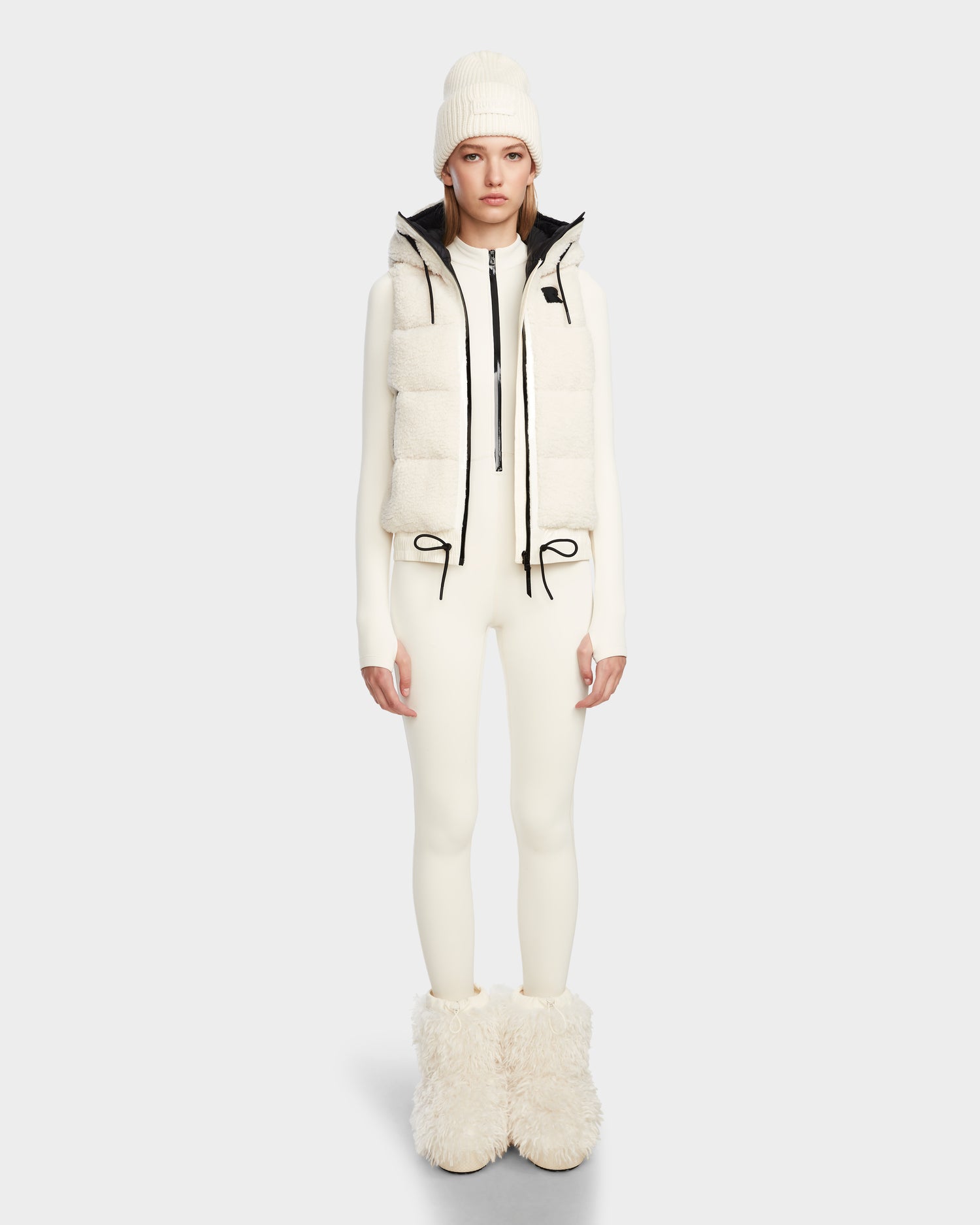 A woman faces forward in a Rudsak RAQUEL S - 8124526 Iceland all-white outfit—knit beanie, teddy ski vest with black drawstrings, fitted long-sleeve suit, and large fuzzy boots—set against a plain light gray background.