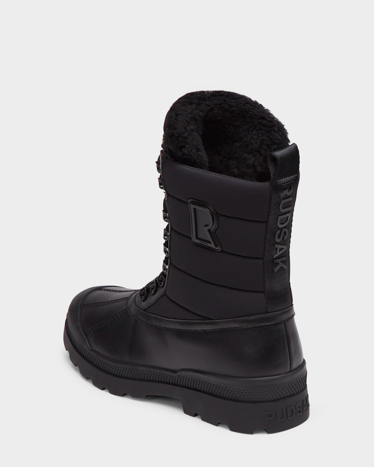 The Rudsak REAGAN - 6224501 Black men’s winter boot features a thick rubber sole, lace-up front, faux fur lining, side branding, and back pull tab. Shown on a plain white background.