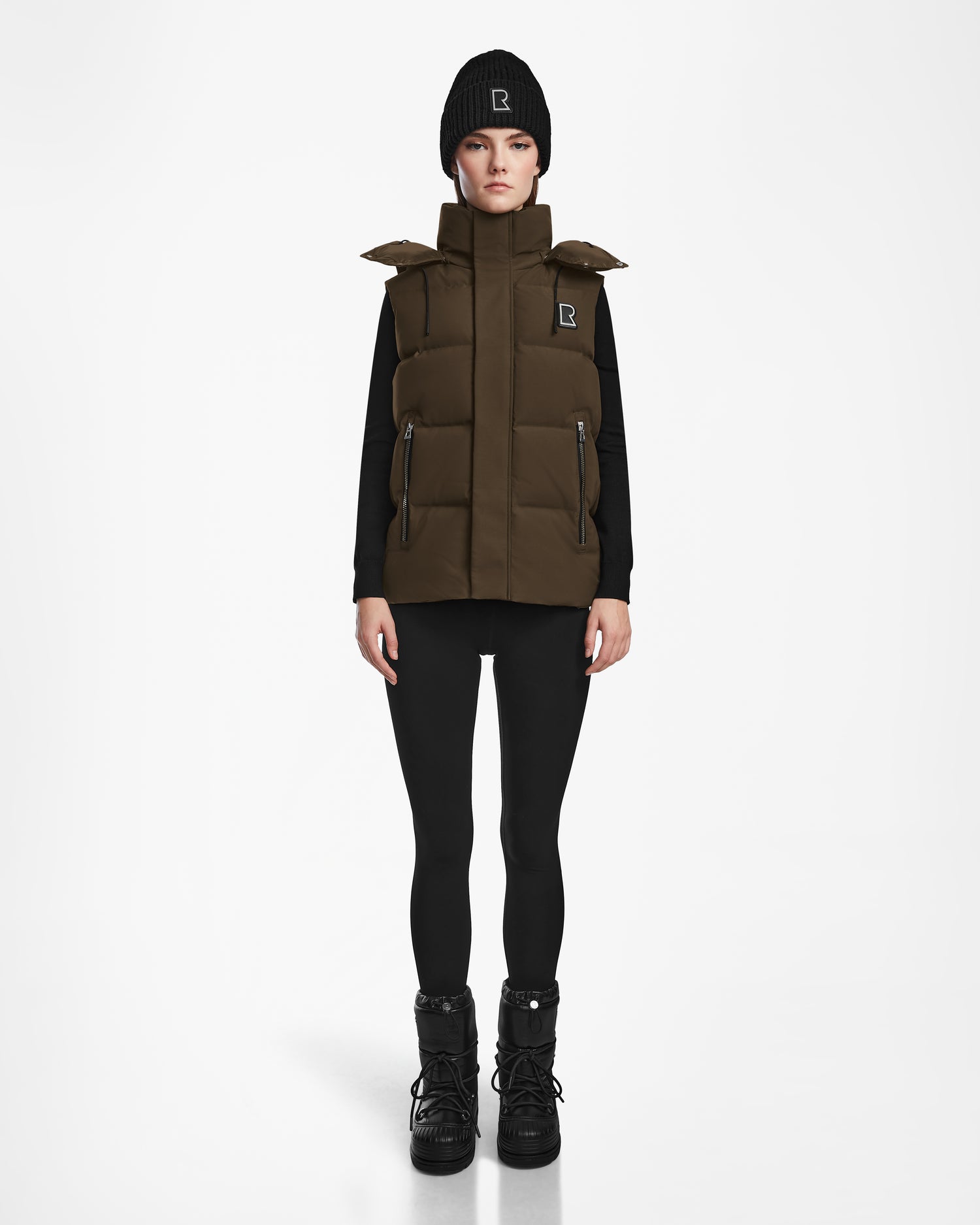 A person wears the Rudsak REN M - 9123512 Army W puffer vest with a high collar over a black long-sleeve top, black leggings, boots, and a beanie, standing facing forward against a plain white background.