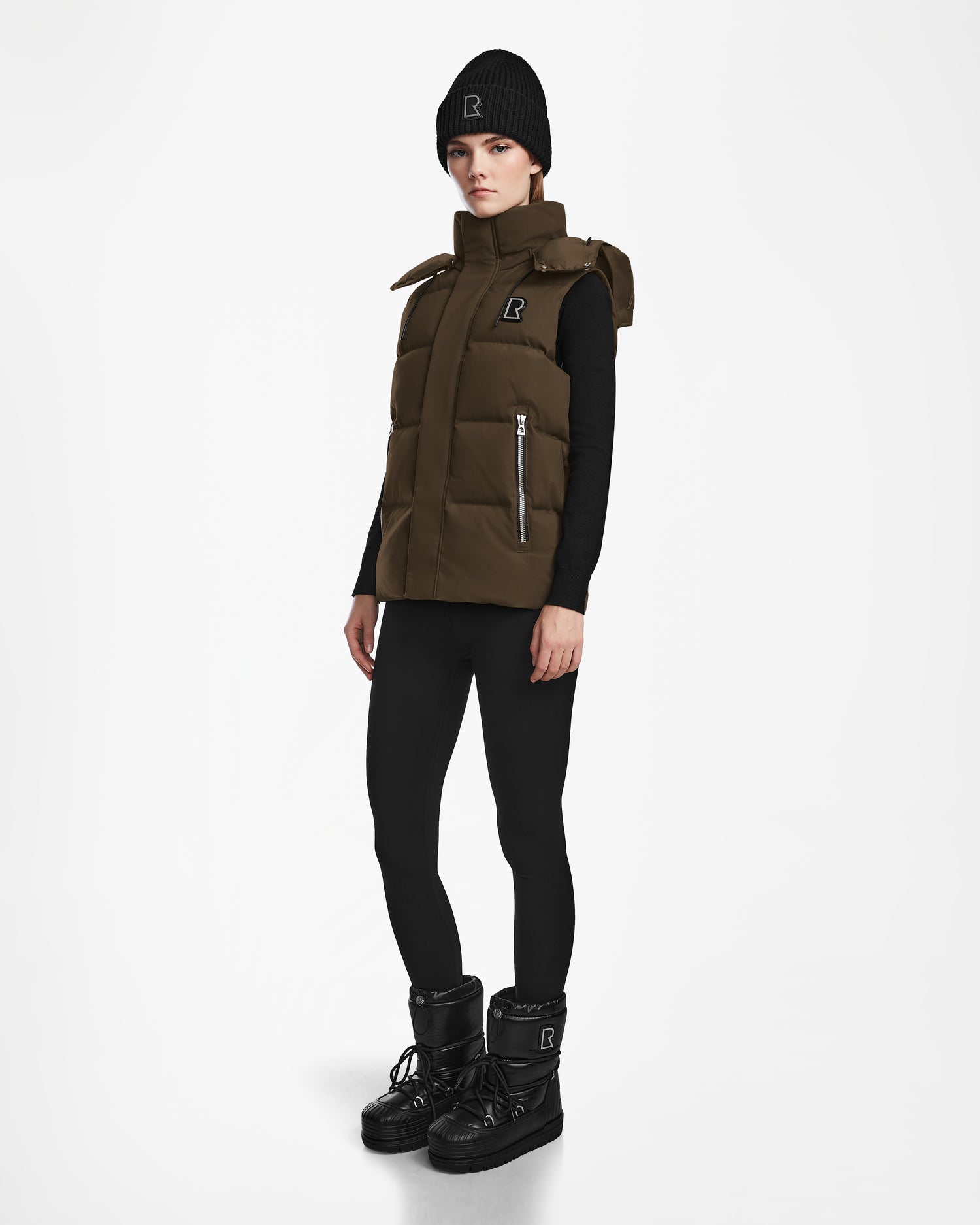 A person wearing the Rudsak REN M - 9123512 Army W weather-resistant brown puffer vest, black beanie, black long-sleeve top, black leggings, and black platform winter boots stands against a plain white background.