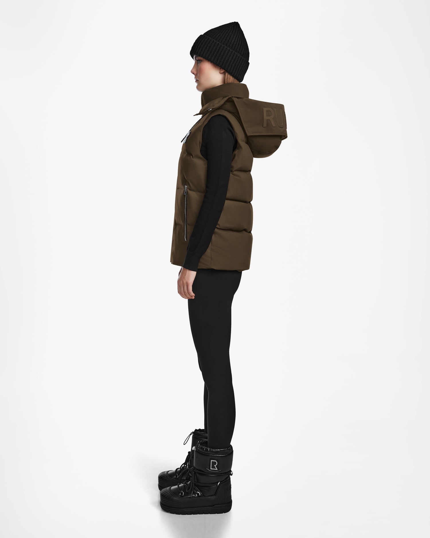 A person stands in profile against a white background, wearing the Rudsak REN M - 9123512 Army W weather-resistant black beanie, a brown puffer vest with hood, black long-sleeve top and leggings, and sturdy black winter boots.