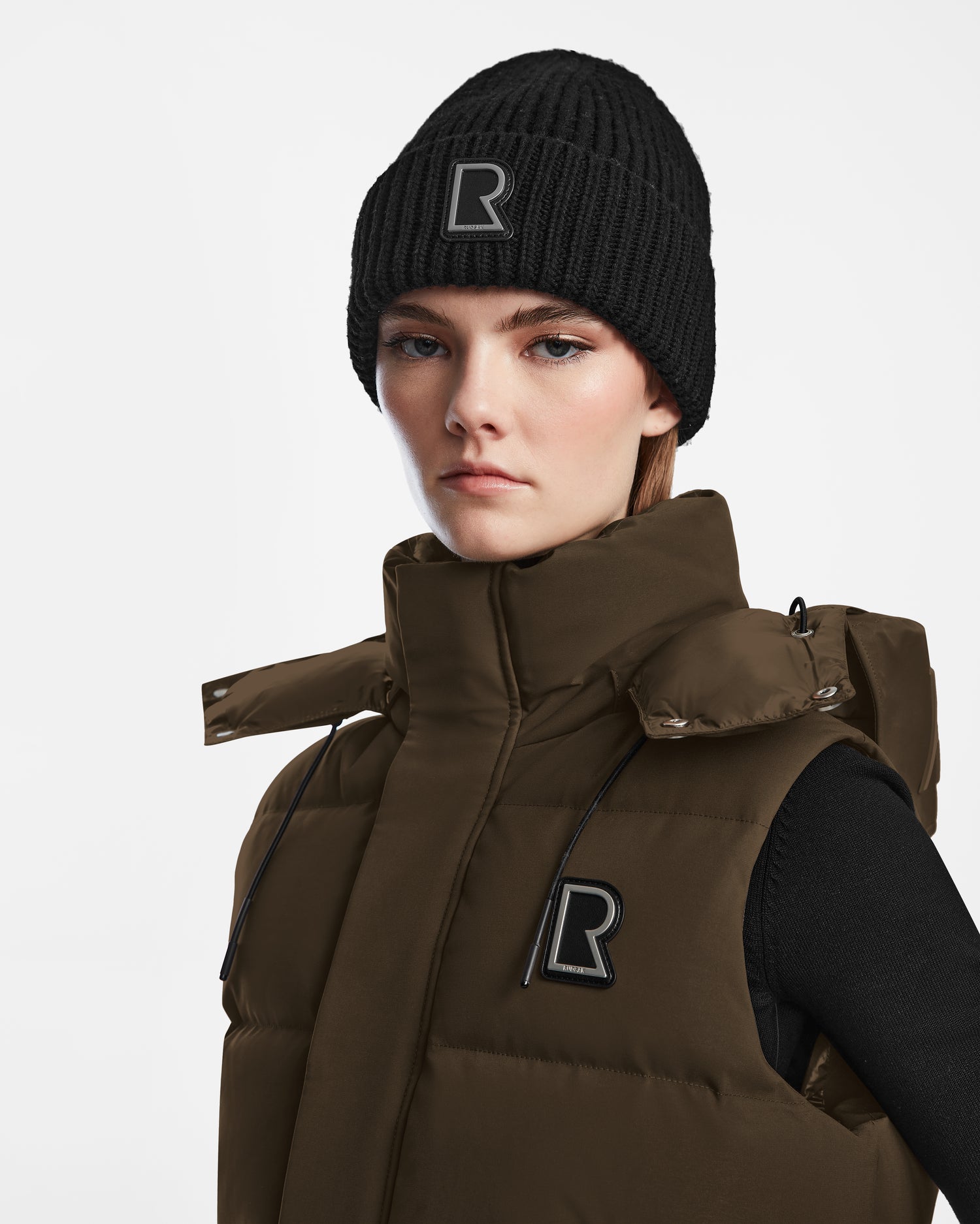 A woman in the Rudsak REN M - 9123512 Army W dark olive, weather-resistant puffer vest with a high collar and R logo, plus a black knit beanie, stands against a white background with a neutral expression and light makeup.
