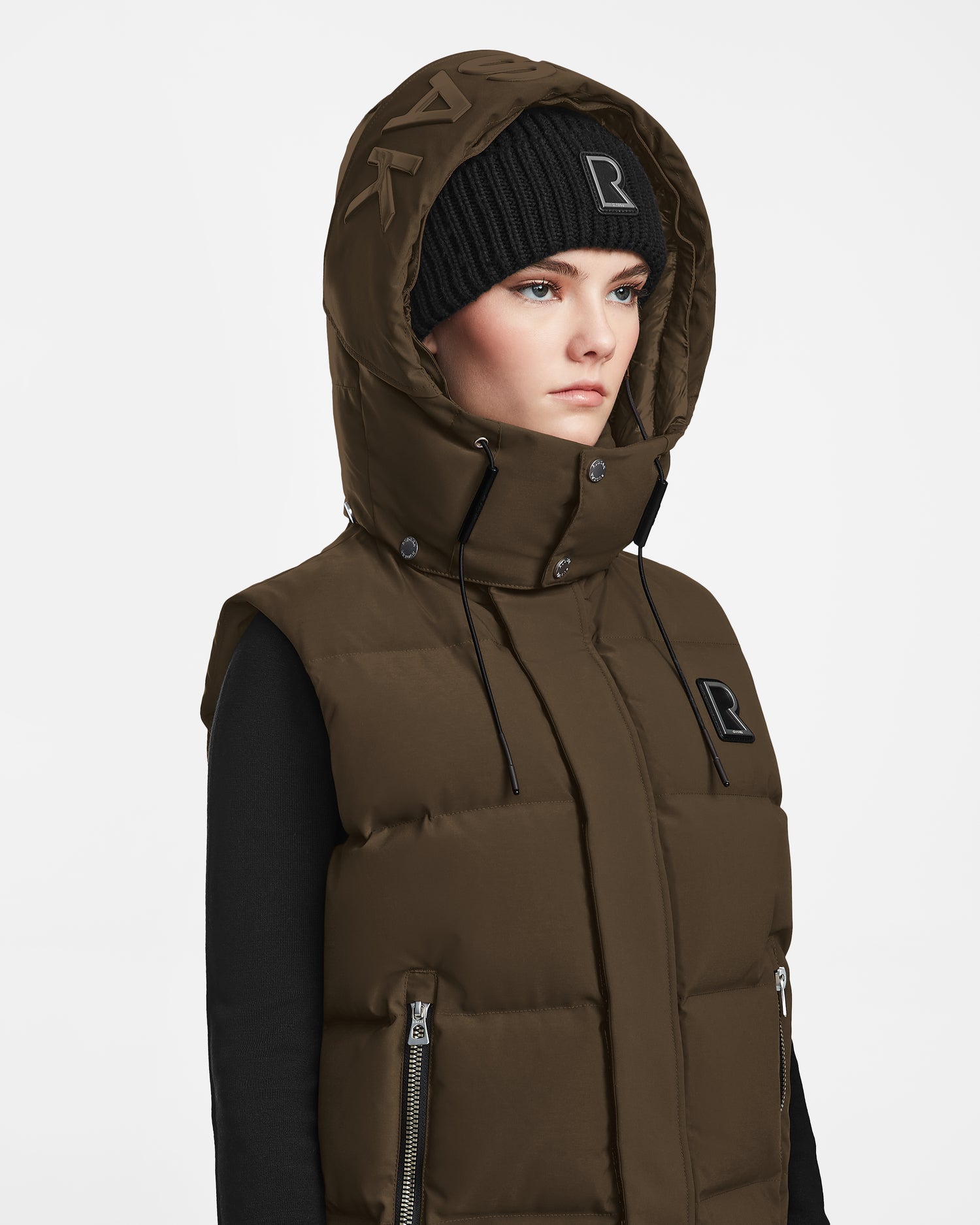 A person wears the Rudsak REN M - 9123512 Army W brown weather-resistant puffer vest with hood, zippers, patches, and RDS-certified down over a black long-sleeve top and black knit beanie, gazing forward against a white background.