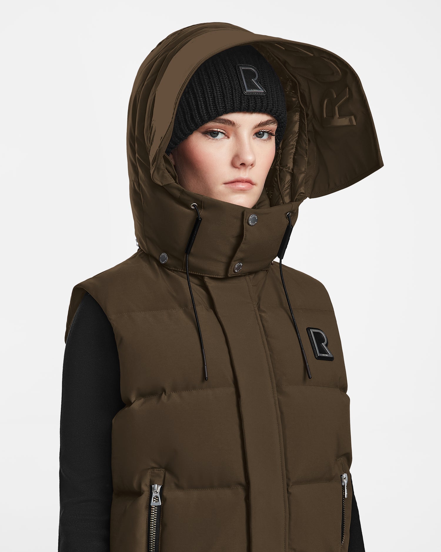A person poses against a white background wearing Rudsaks REN M - 9123512 Army W weather-resistant brown sleeveless puffer vest with a large hood and a black beanie featuring an R.