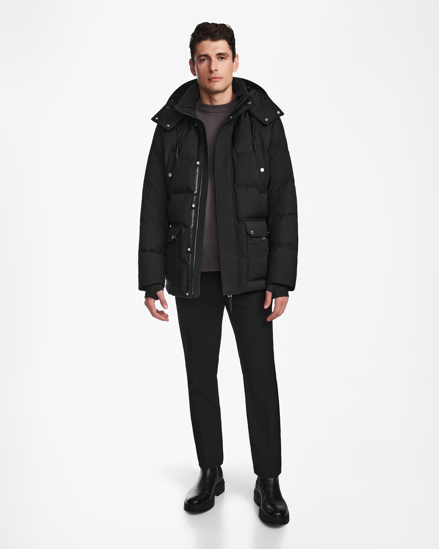 A man faces forward against a white background, wearing the Rudsak RICKY - M123502 Black parka over a dark sweater, black pants, and black boots. His posture is relaxed and his expression neutral.