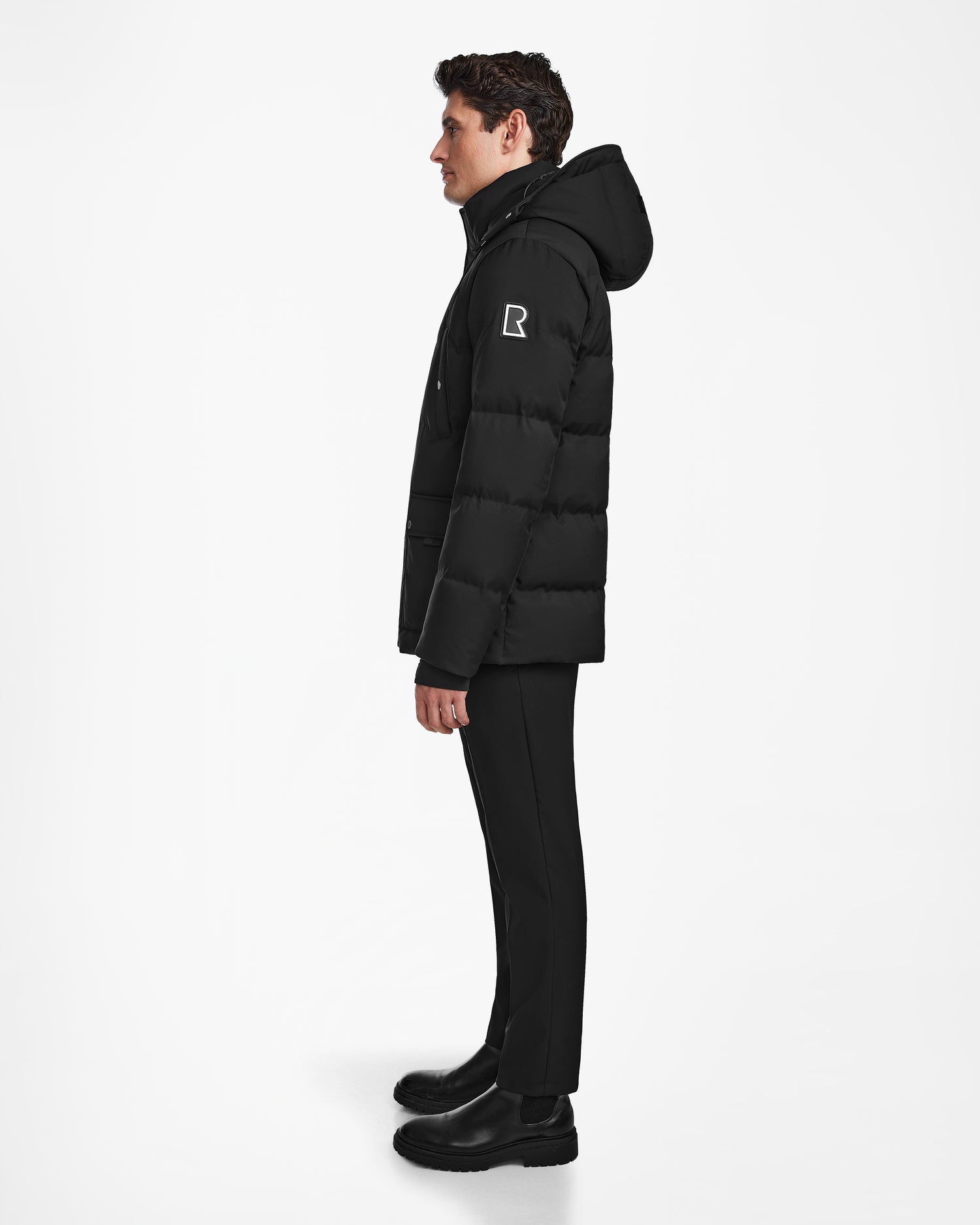 A person stands in profile against a white background wearing the Rudsak RICKY - M123502 Black parka, black pants, and black boots. The winter parka features a white R logo on the upper sleeve.