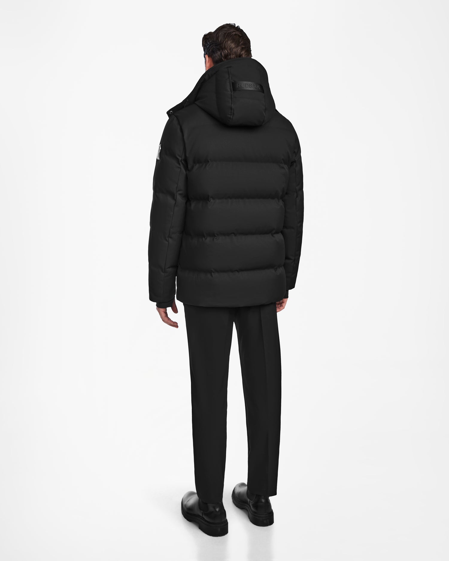 A person stands facing away, wearing the Rudsak RICKY - M123502 Black parka with a hood, paired with black pants and shoes, against a plain white background.