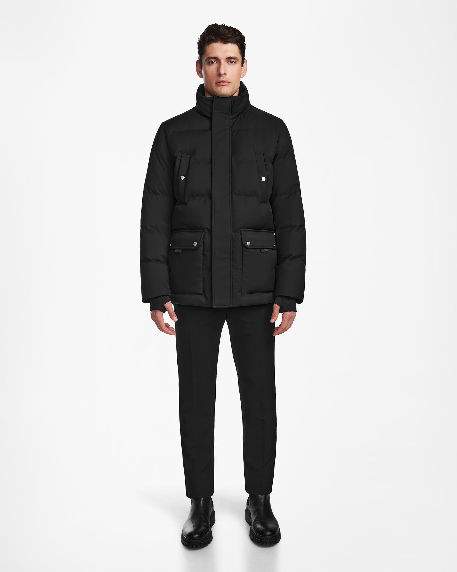 A man stands facing forward wearing the Rudsak RICKY - M123502 Black men's quilted down parka with large pockets, black pants, and black boots against a plain white background.
