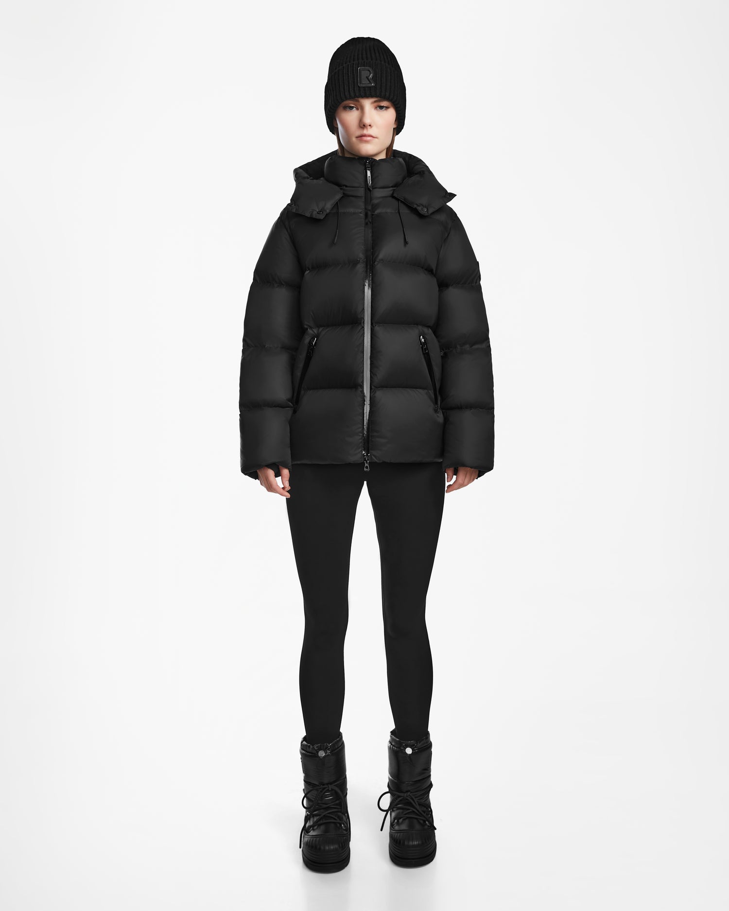 A person faces forward against a plain white background, wearing Rudsak's RITA M - L124537 Black weather-resistant puffer jacket, paired with black leggings, black winter boots, and a black knit beanie.