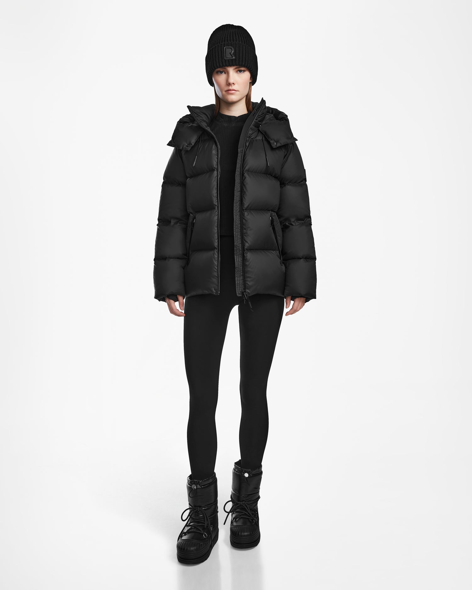 A person faces forward against a white background, wearing the Rudsak RITA M - L124537 Black weather-resistant puffer jacket, styled with a black beanie, black leggings, and black boots.