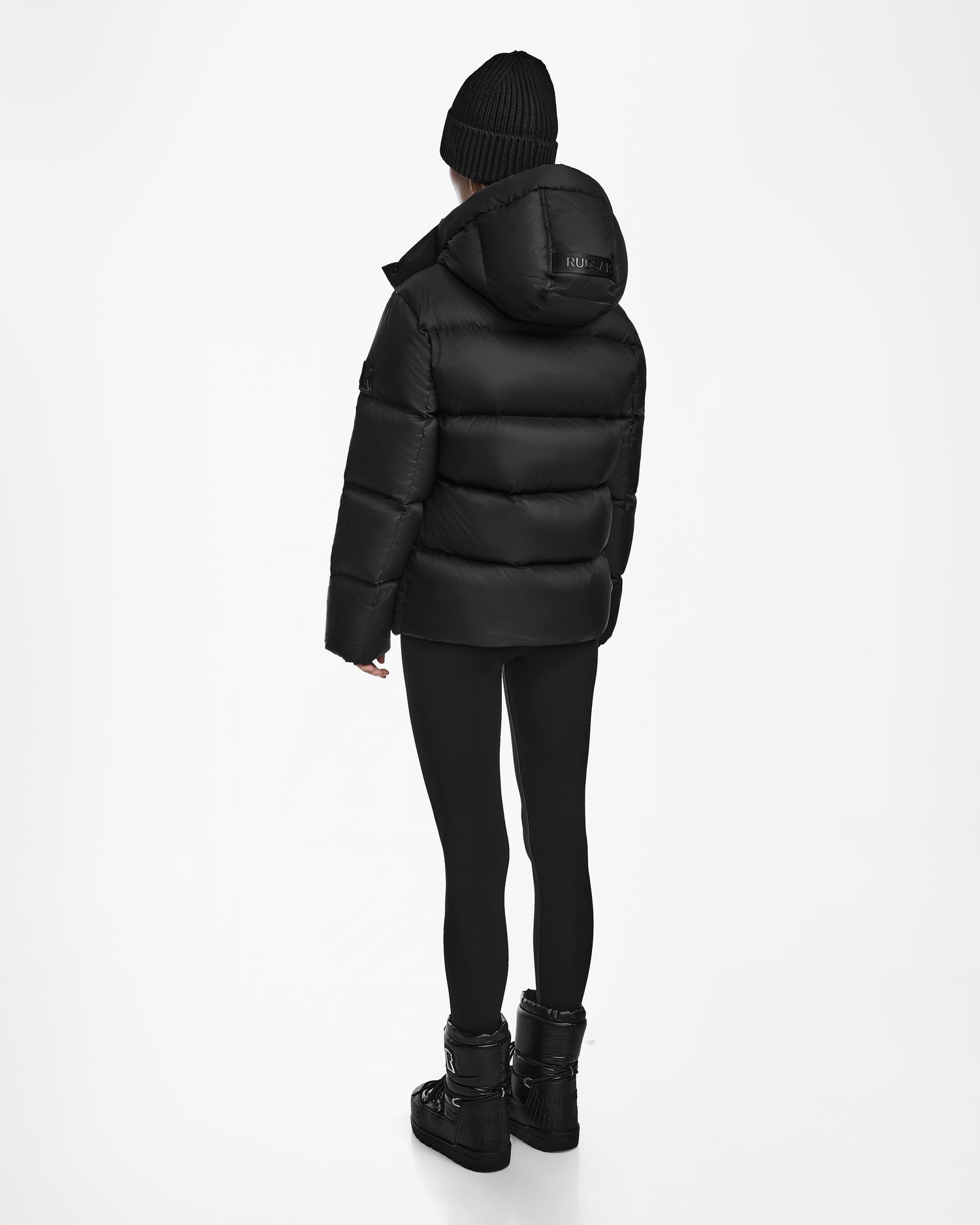 A person wearing the Rudsak RITA M - L124537 Black weather-resistant puffer, paired with a beanie, leggings, and boots, stands facing away on a white background.