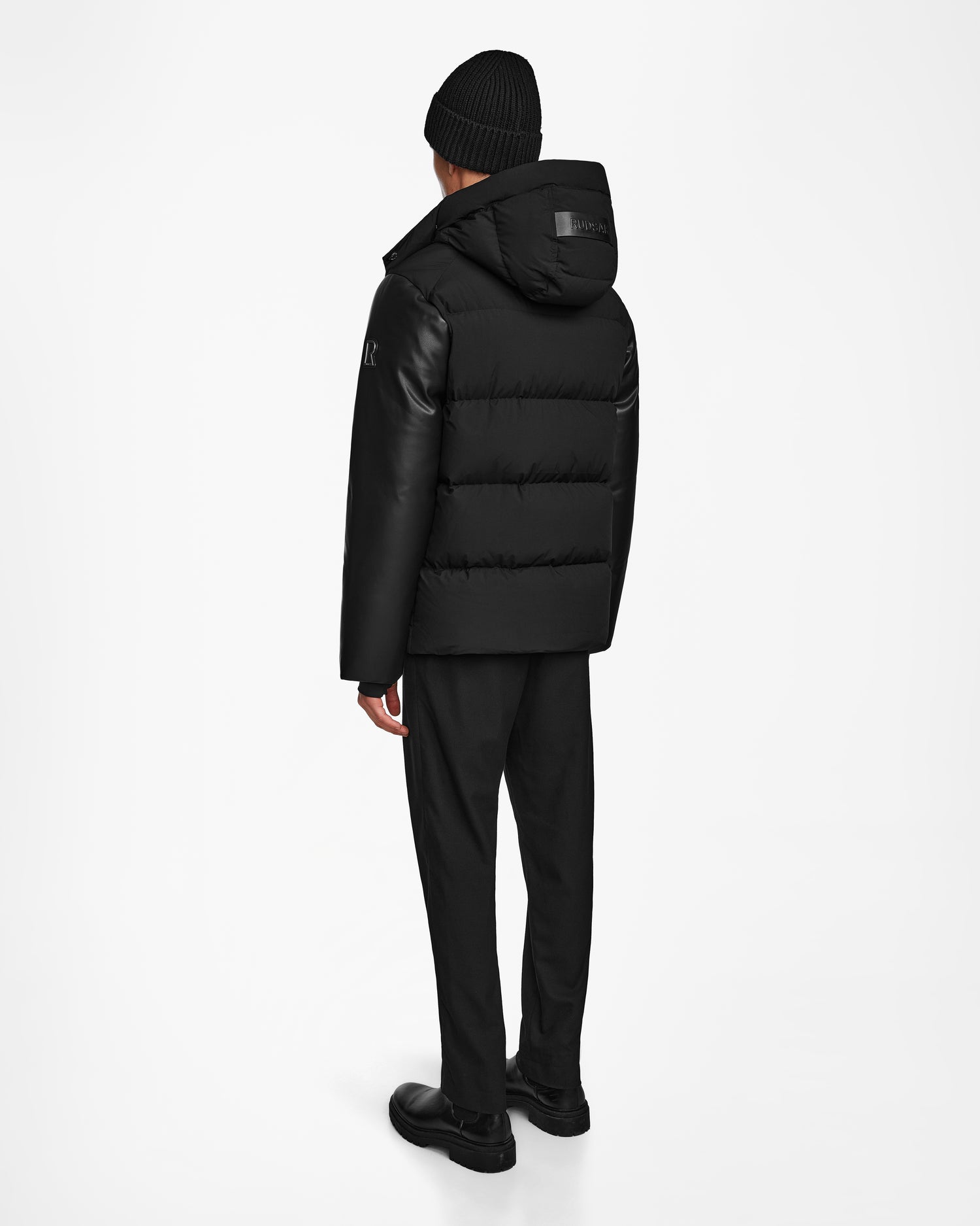 A person stands facing away, wearing the Rudsak RONNY M - M124513 Black hooded puffer jacket made from recycled materials, paired with black pants and shoes against a plain white background.