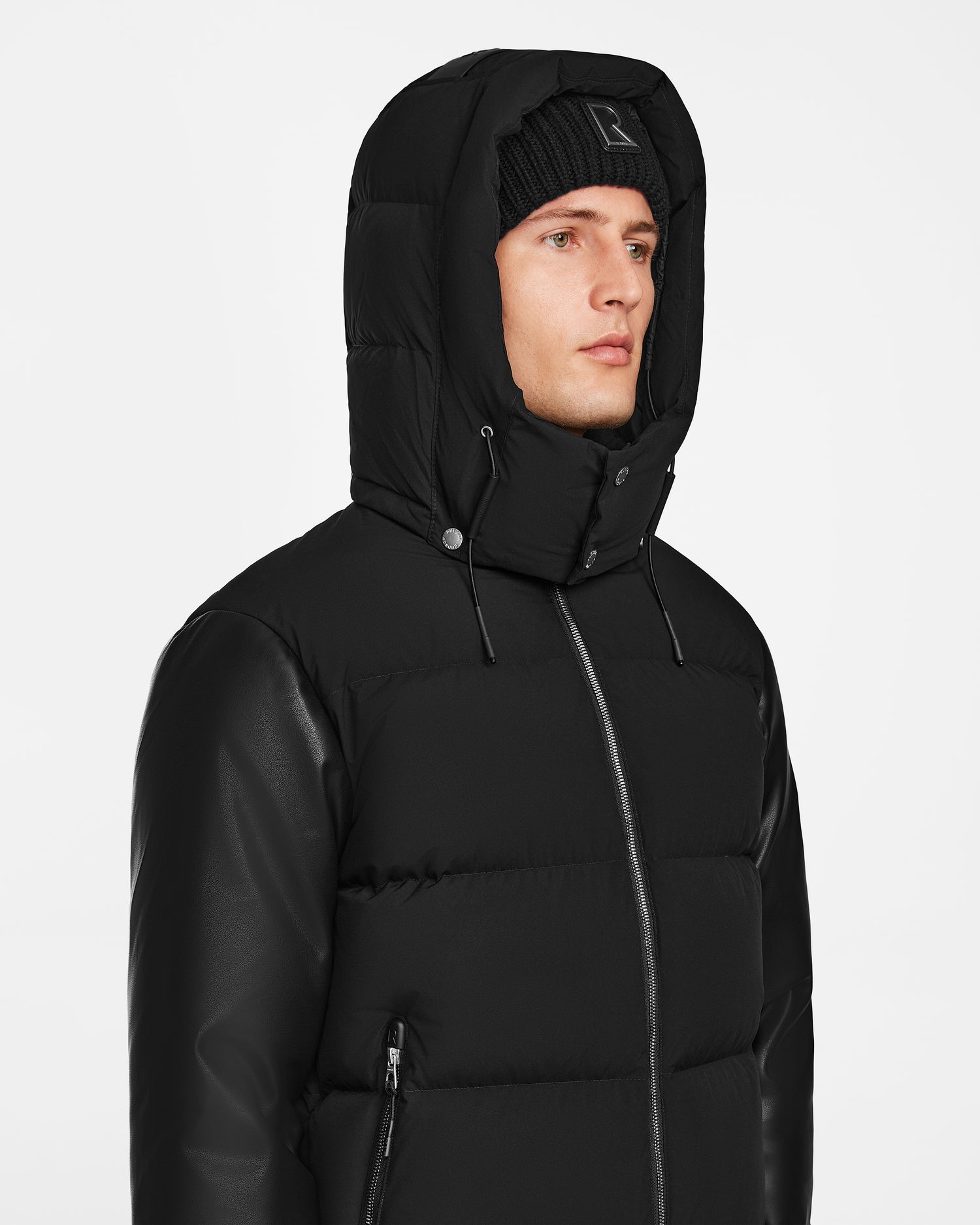 A person wearing the Rudsak RONNY M - M124513 Black puffer jacket with a hood and a black knit beanie, looking slightly to the side against a plain white background.