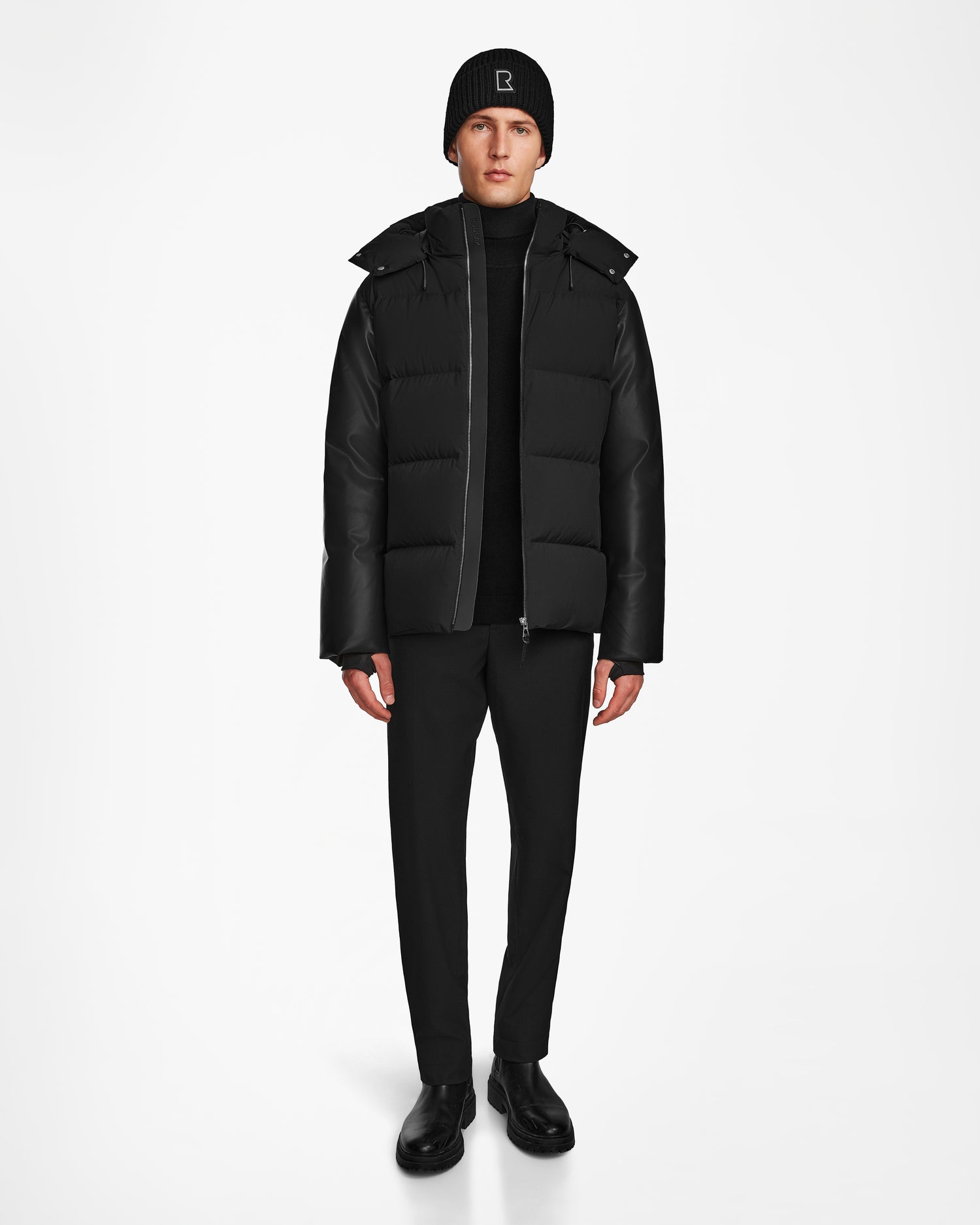 A person wears the Rudsak RONNY M - M124513 Black puffer jacket, paired with black pants, boots, and a beanie, standing against a plain white background facing forward with arms at their sides.