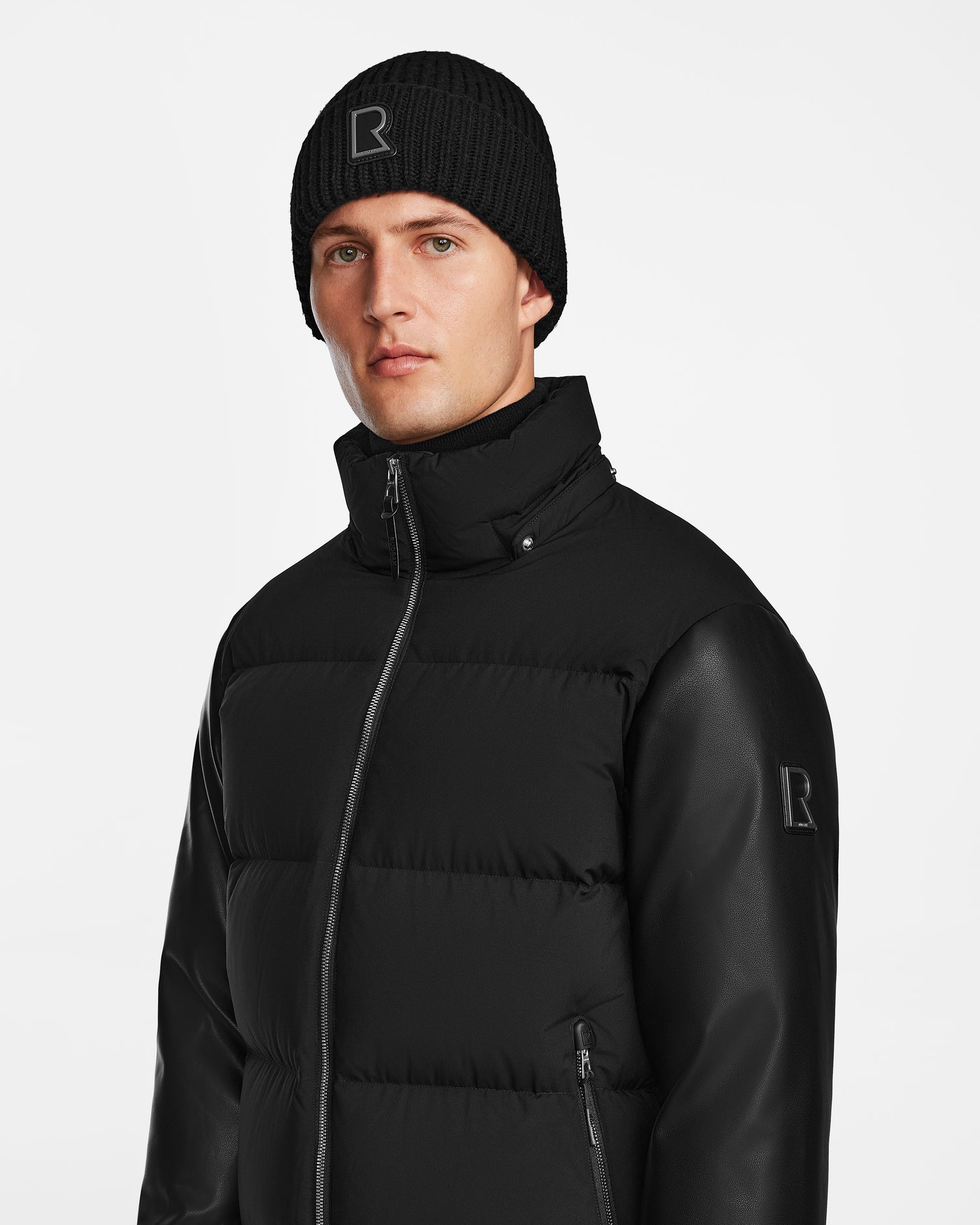 A man stands against a white background wearing the Rudsak RONNY M - M124513 Black quilted down jacket with leather sleeves and matching black knit beanie, both featuring the signature R logo—an essential modern winter look.