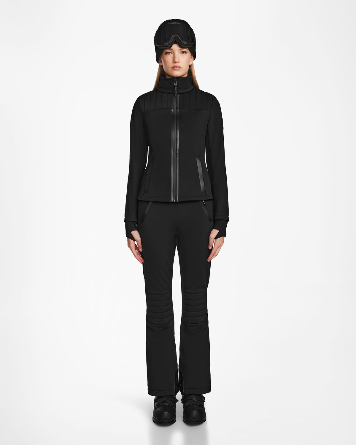 A woman faces forward wearing the Rudsak ROSALIE - 8124784 Black fitted ski jacket with matching black pants, boots, and a black winter hat, set against a plain white background.