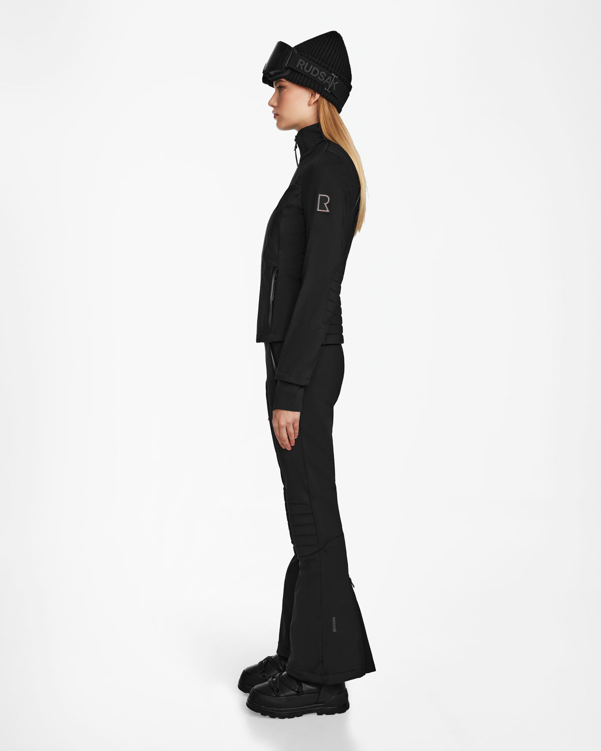 A woman stands in profile against a plain white background, wearing the Rudsak ROSALIE - 8124784 Black fitted ski jacket, flared pants, winter boots, and a knit beanie hat with RUDSAK written on it.