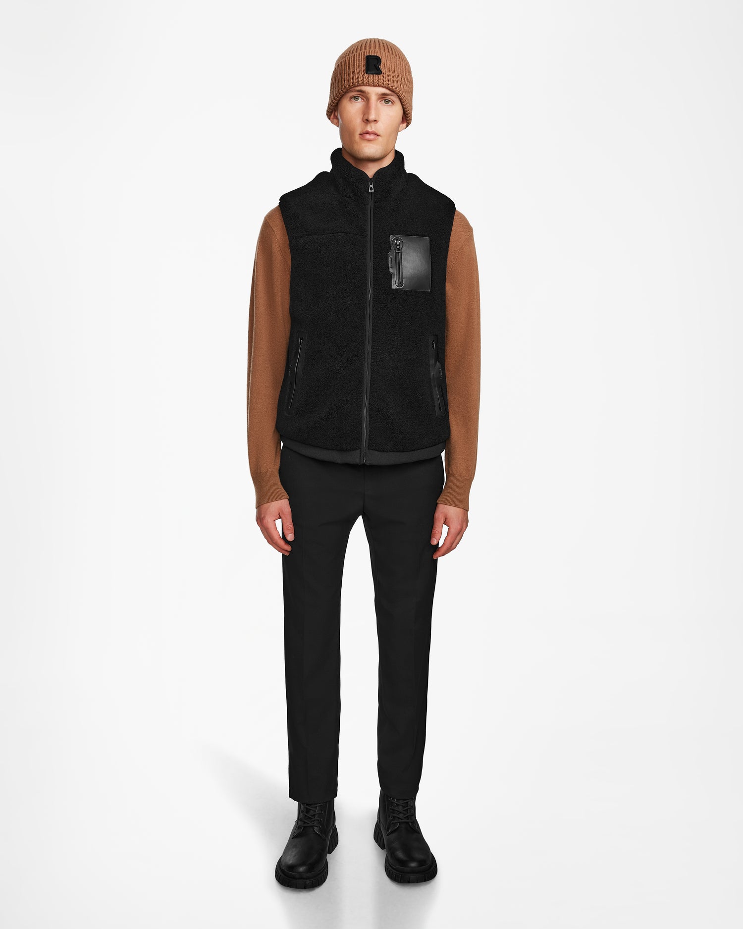 A person stands facing forward, wearing the Rudsak RUMMY - U524502 Black M unisex vest with a chest pocket, paired with a brown knit beanie, brown long-sleeve shirt, black pants, and black boots against a plain white background.