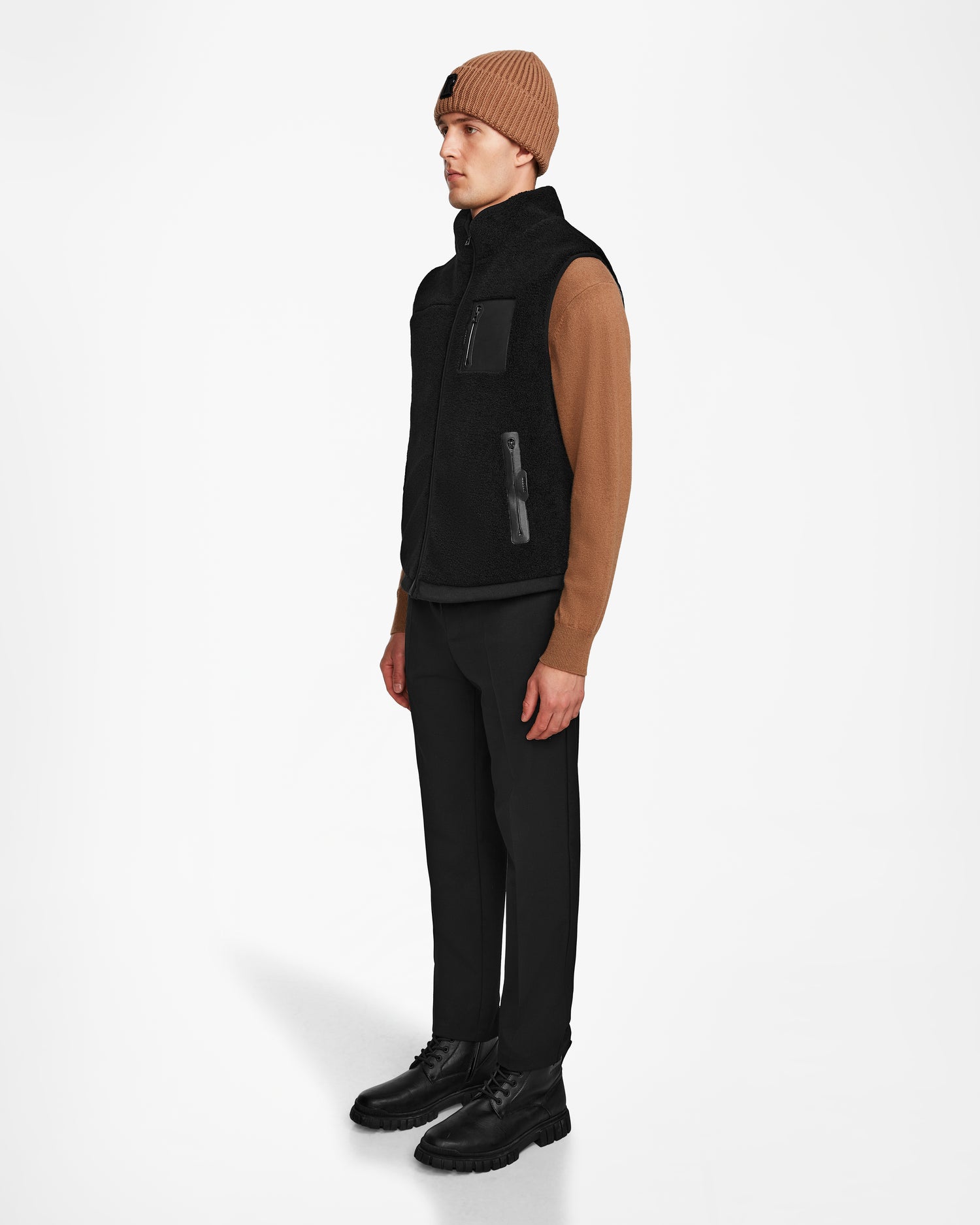 A person in a brown beanie, tan sweater, Rudsaks RUMMY - U524502 Black M unisex vest with pockets, black pants, and black lace-up boots stands facing left against a plain white background.