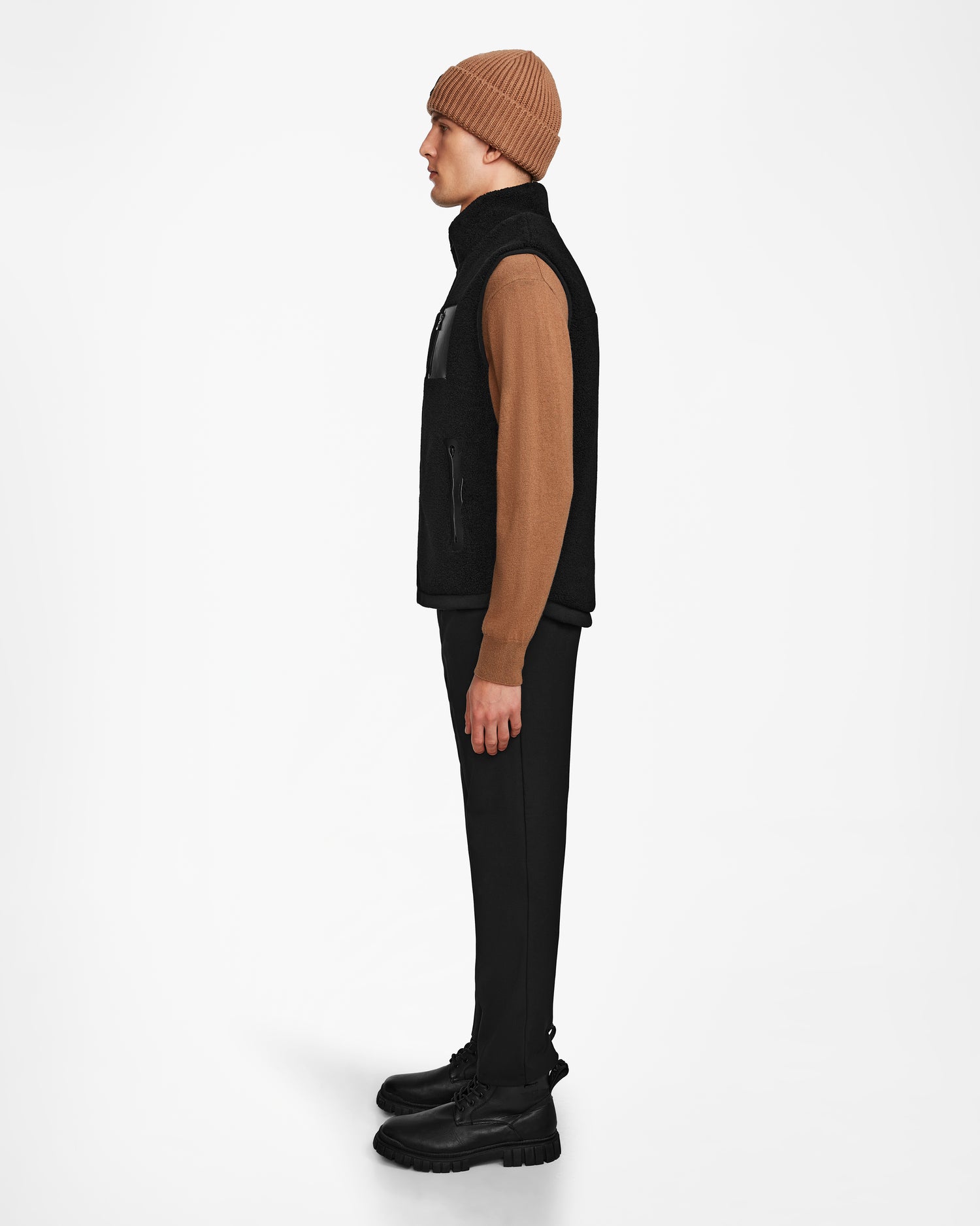 A person stands in profile against a white background, wearing the Rudsak RUMMY - U524502 Black M vest, paired with a brown beanie, tan sweater, black pants, and black boots.
