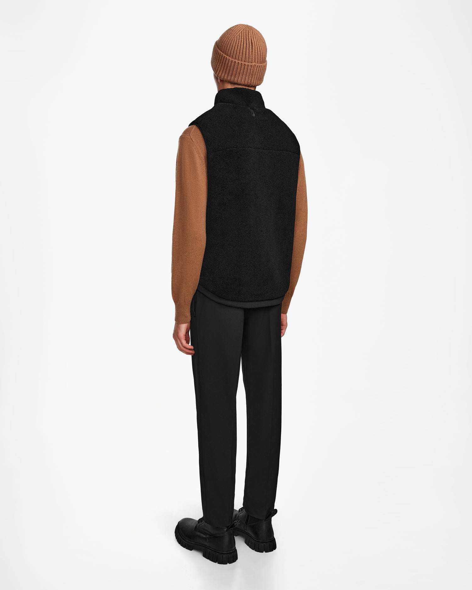 A person stands facing away, wearing the Rudsak RUMMY - U524502 Black M vest, paired with a brown knit beanie, brown long-sleeve top, black pants, and black shoes against a plain white background.