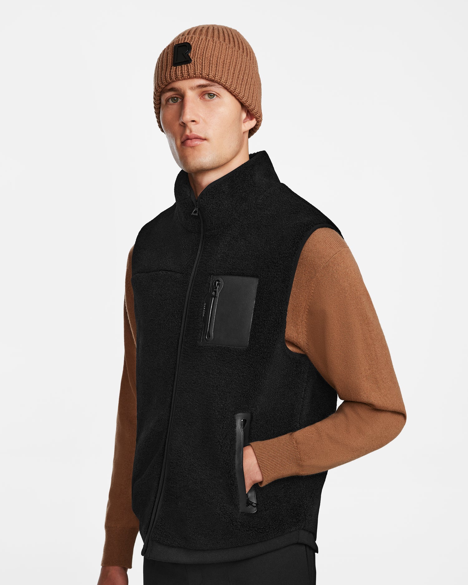 A man wears a brown knit beanie, a brown long-sleeve shirt, and the Rudsak RUMMY - U524502 Black M zip-up polar fleece vest with zippered pockets, standing against a plain white background.