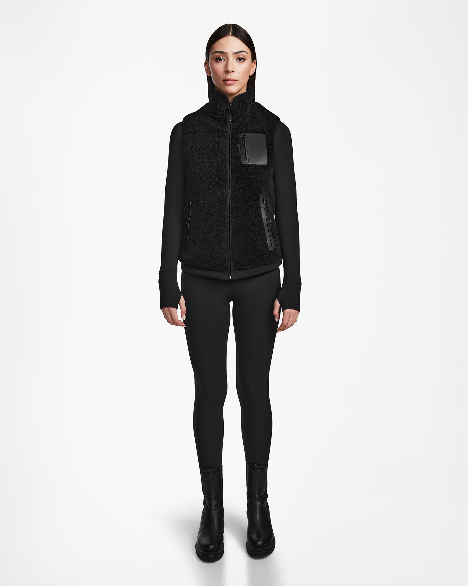 A person stands facing forward, dressed in Rudsak’s RUMMY - U524502 Black W, paired with a black top, leggings, and boots, against a plain white background.