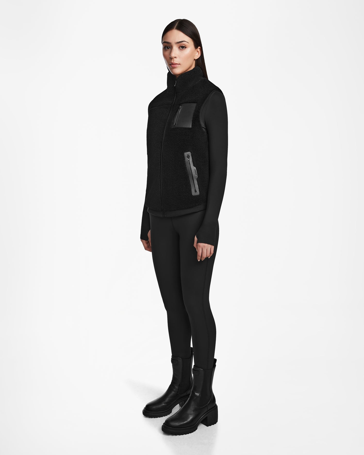 A woman faces forward against a white background, wearing the Rudsak RUMMY - U524502 Black W polar fleece vest over a fitted black top, black leggings, and ankle boots. She has zippered pockets and maintains a neutral expression.