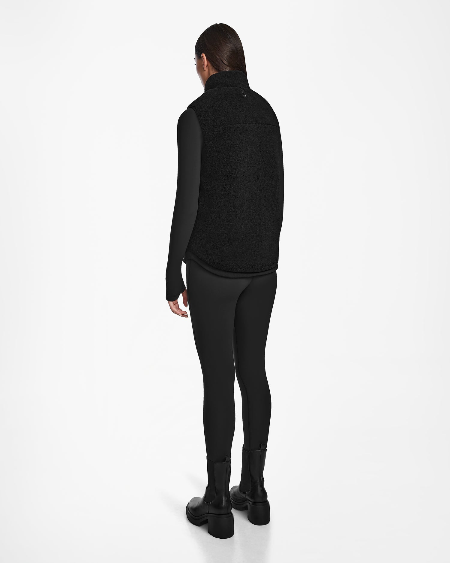 A person with long brown hair faces away, wearing the Rudsak RUMMY - U524502 Black W unisex zip-up vest, long-sleeve top, leggings, and boots against a plain white background.